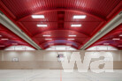 Der Innenraum der Sporthalle definiert sich über das Dachtragwerk. Die rote Dachkonstruktion bildet einenbewussten Kontrast zu den Holzflächen der Prallwände und des Sportbodens